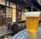 Plastic cup of pale beer with a foam head on a weathered blue table outdoors, rustic wooden building in the background.