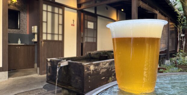 Plastic cup of pale beer with a foam head on a weathered blue table outdoors, rustic wooden building in the background.