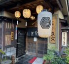 Traditional Japanese storefront with hanging lanterns and wooden lattice doors, vertical signage in Japanese on the walls.