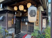 Traditional Japanese storefront with hanging lanterns and wooden lattice doors, vertical signage in Japanese on the walls.
