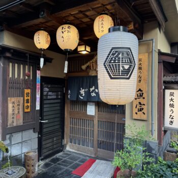 Traditional Japanese storefront with hanging lanterns and wooden lattice doors, vertical signage in Japanese on the walls.