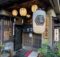 Traditional Japanese storefront with hanging lanterns and wooden lattice doors, vertical signage in Japanese on the walls.