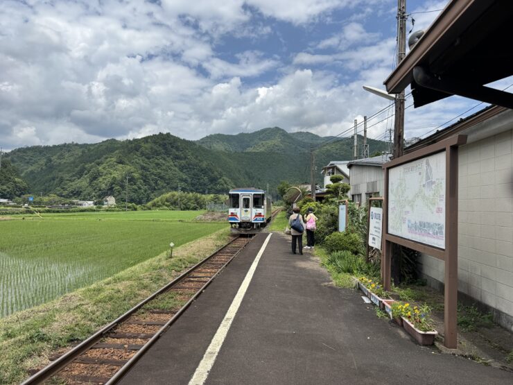 Two people stand on a rural train platform beside tracks, with a small blue-and-white train and green hills in the distance under a cloudy sky.
