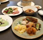 Assorted Asian dishes on a table: noodles, dumplings, fried chicken, greens, and a creamy yellow sauce on a plate with a drink nearby.