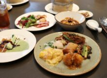 Assorted Asian dishes on a table: noodles, dumplings, fried chicken, greens, and a creamy yellow sauce on a plate with a drink nearby.
