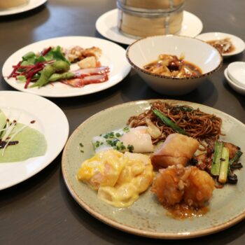 Assorted Asian dishes on a table: noodles, dumplings, fried chicken, greens, and a creamy yellow sauce on a plate with a drink nearby.