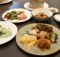 Assorted Asian dishes on a table: noodles, dumplings, fried chicken, greens, and a creamy yellow sauce on a plate with a drink nearby.