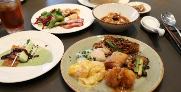 Assorted Asian dishes on a table: noodles, dumplings, fried chicken, greens, and a creamy yellow sauce on a plate with a drink nearby.