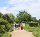 Group of five visitors walking along a gravel garden path, surrounded by trees and lush greenery on a sunny day.
