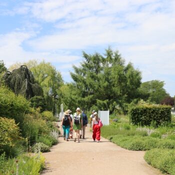 Group of five visitors walking along a gravel garden path, surrounded by trees and lush greenery on a sunny day.
