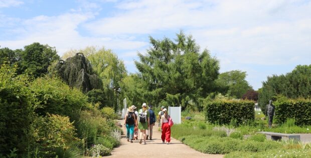 Group of five visitors walking along a gravel garden path, surrounded by trees and lush greenery on a sunny day.