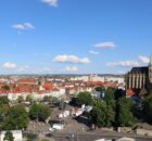 Aerial city view with red-tiled roofs, a Gothic cathedral with green spires on the right, and a busy square below under a blue sky.