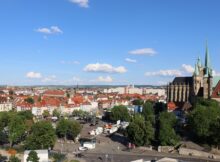 Aerial city view with red-tiled roofs, a Gothic cathedral with green spires on the right, and a busy square below under a blue sky.