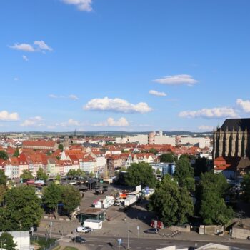 Aerial city view with red-tiled roofs, a Gothic cathedral with green spires on the right, and a busy square below under a blue sky.