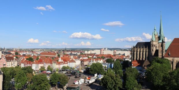 Aerial city view with red-tiled roofs, a Gothic cathedral with green spires on the right, and a busy square below under a blue sky.