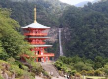 Red, three-story pagoda temple set in a lush green valley with a waterfall in the background.