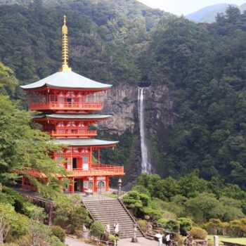 Red, three-story pagoda temple set in a lush green valley with a waterfall in the background.
