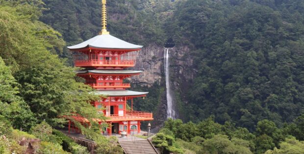 Red, three-story pagoda temple set in a lush green valley with a waterfall in the background.