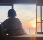 Back view of a person in a patterned robe sitting on a window seat, watching a sunset over the water outside.