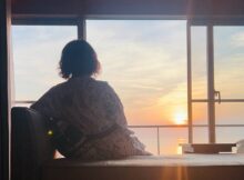 Back view of a person in a patterned robe sitting on a window seat, watching a sunset over the water outside.