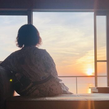 Back view of a person in a patterned robe sitting on a window seat, watching a sunset over the water outside.