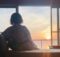 Back view of a person in a patterned robe sitting on a window seat, watching a sunset over the water outside.