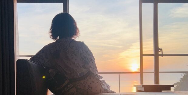 Back view of a person in a patterned robe sitting on a window seat, watching a sunset over the water outside.