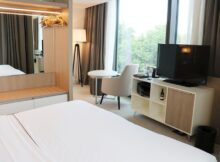 Modern hotel room with a large window, white bed, and a TV on a white media console; seating area includes a round table and an gray chair.