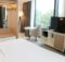 Modern hotel room with a large window, white bed, and a TV on a white media console; seating area includes a round table and an gray chair.